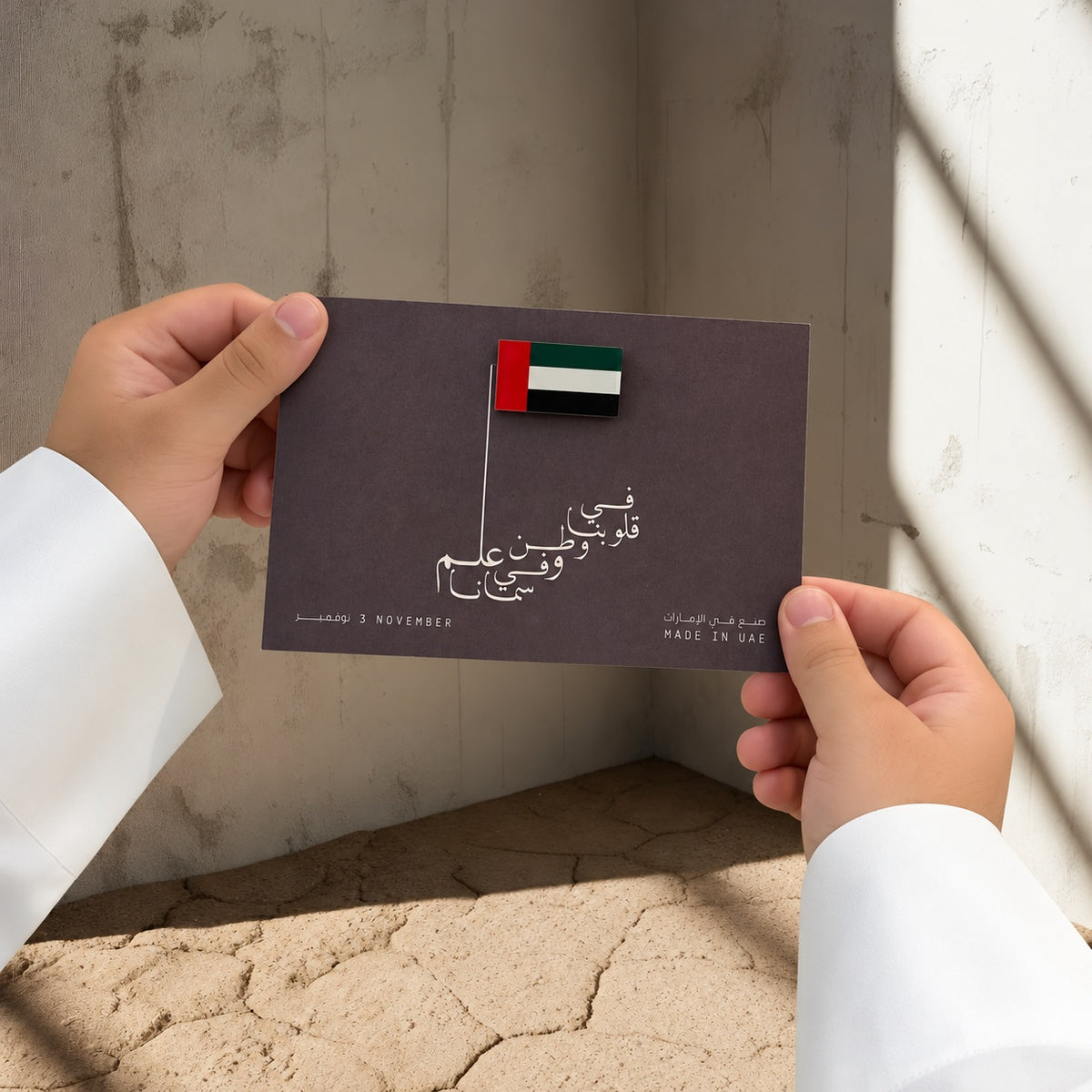يدان بكمّي كندورة بيضاء تحملان بروش علم الإمارات على بطاقة داكنة بخط عربي أبيض وسارية رفيعة.