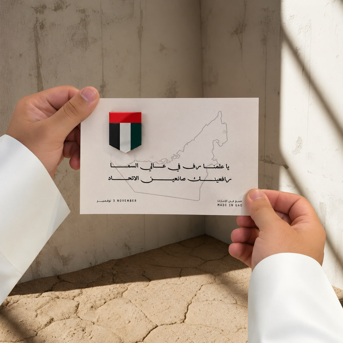 يدان بكمّي كندورة بيضاء تحملان بروش درع بألوان علم الإمارات على بطاقة بخطوط خريطة الإمارات وعبارات عربية.