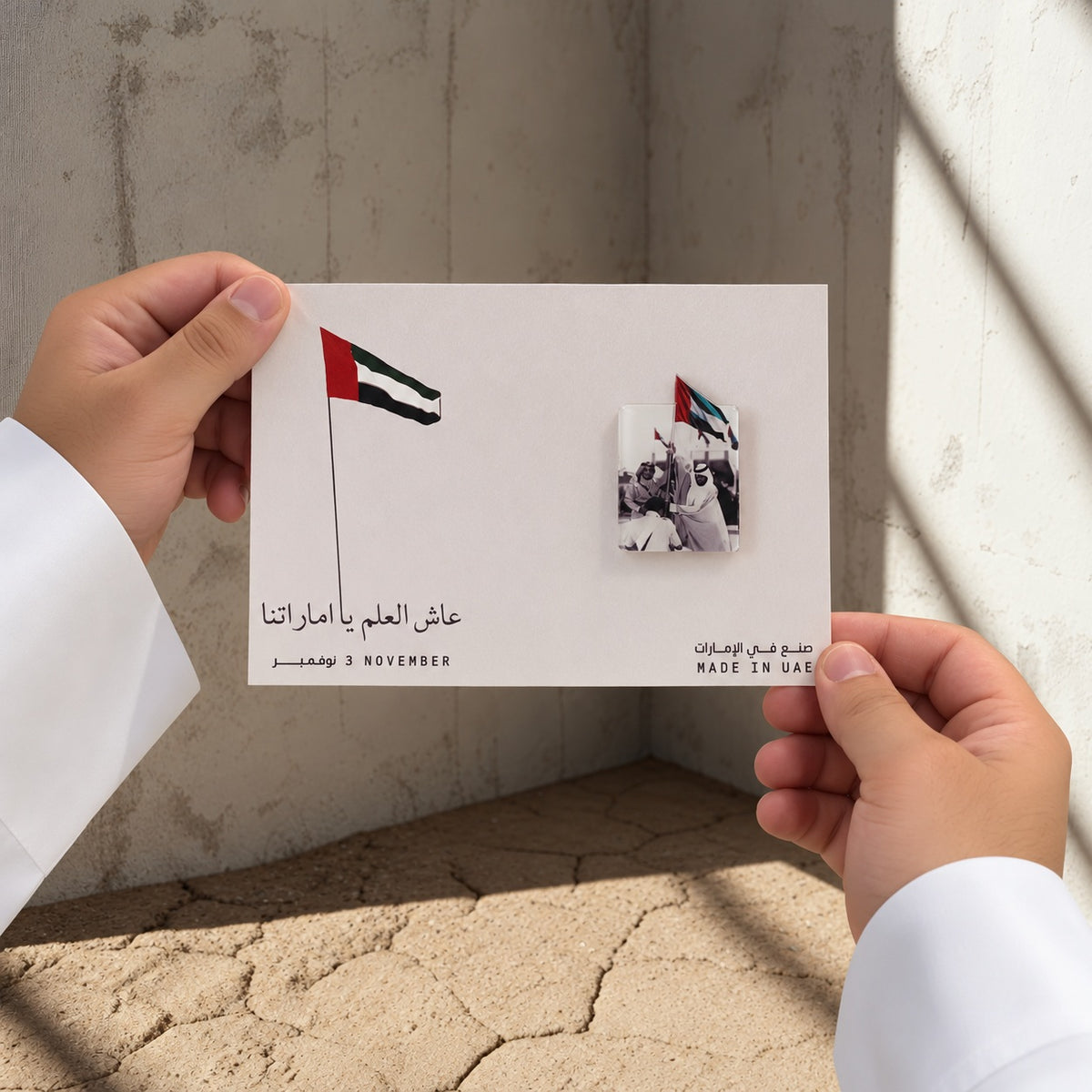 يدان بكمّي كندورة بيضاء تحملان بروشاً مستطيلاً بصورة تراثية وعلم ملون على بطاقة مطبوعة بعلم الإمارات.