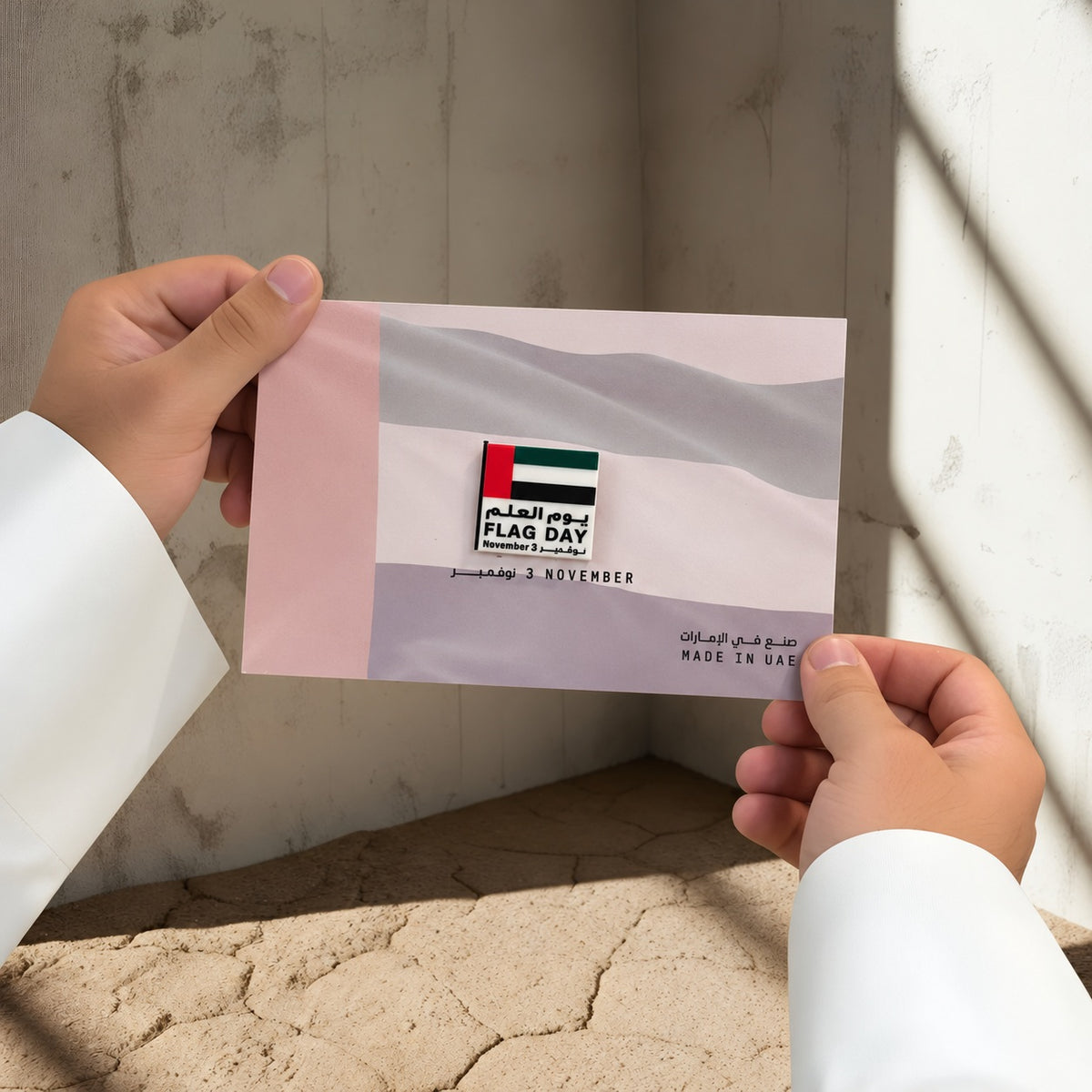 يدان بكمّي كندورة بيضاء تحملان بروش يوم العلم FLAG DAY على بطاقة علم الإمارات بألوان وردية ورمادية هادئة.